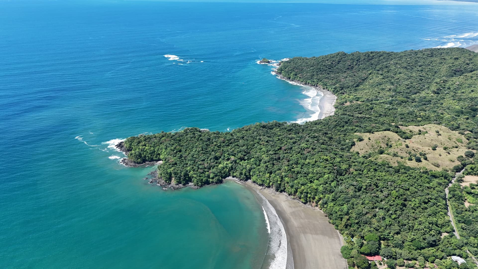 Aerial view of a secluded Pacific shoreline with turquoise water, dark sand beach and forested headlands