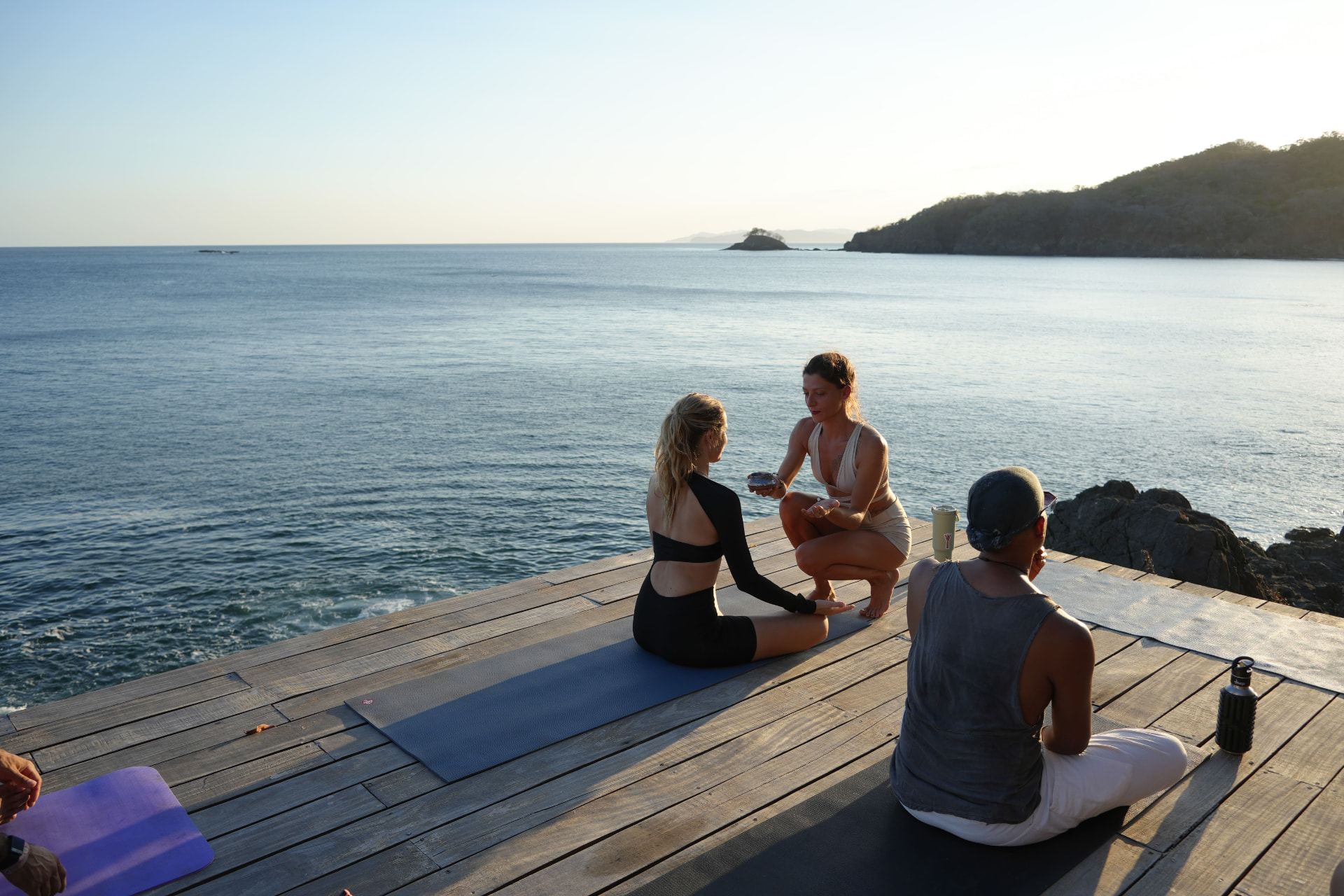 Three people practicing yoga and breathwork on a wooden deck above the Pacific Ocean at sunrise