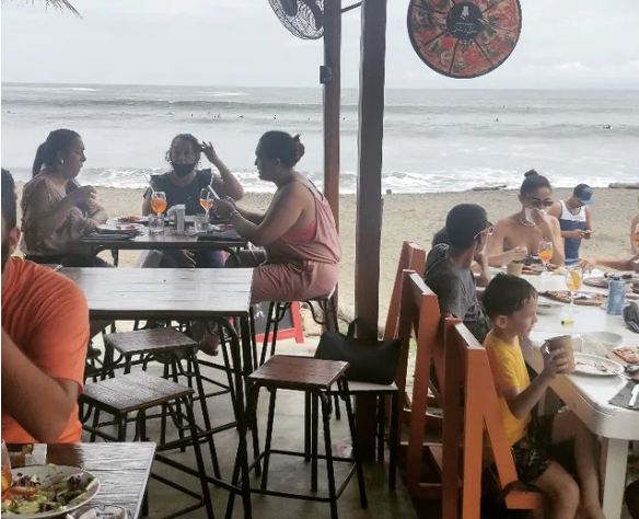 Diners at wooden tables under a palm roof looking out over the surf at Gato Blanco Pizzaria